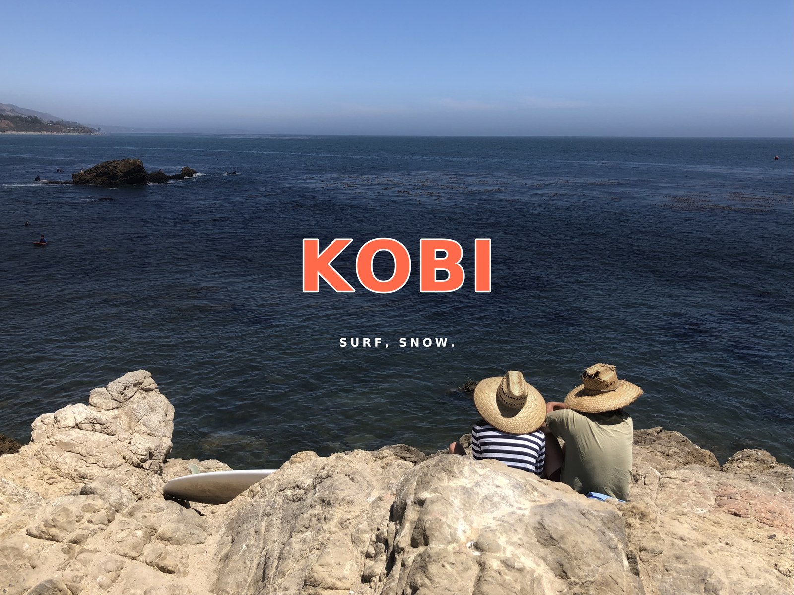 KOBI — Surf, Snow. Two people sitting on coastal rocks in Malibu, looking out at the ocean, with a surfboard beside them.